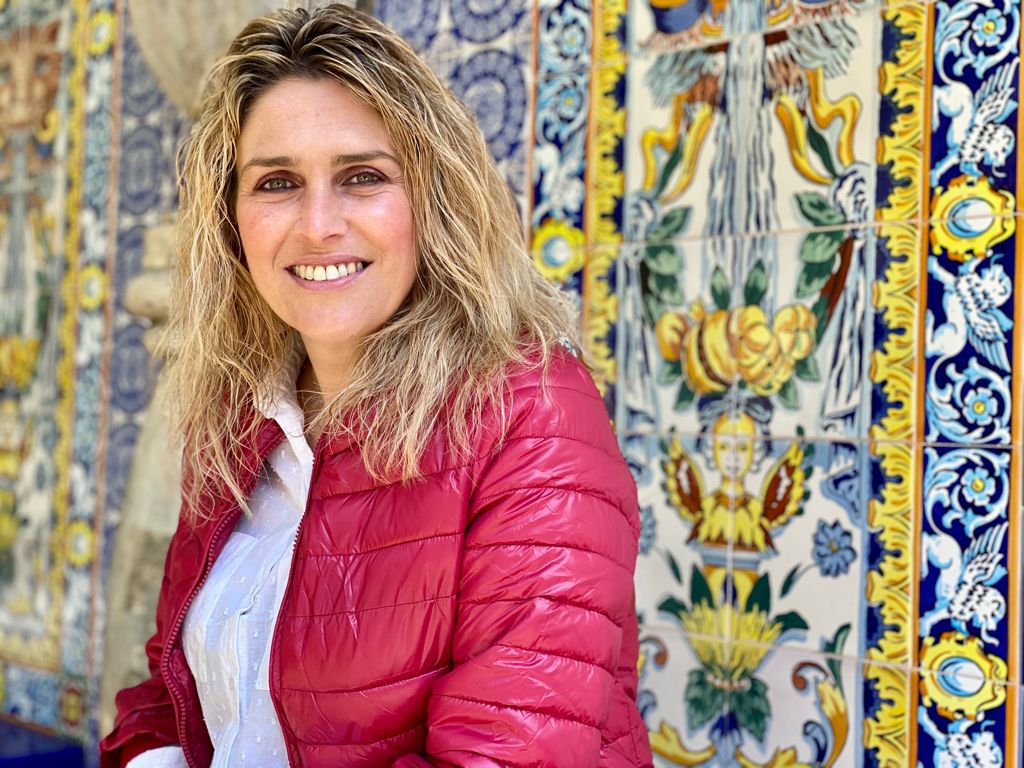 Primer Plano de Marta Barrachina sonriente, con cazadora roja sobre un fondo de azulejos con motivos típicos de la cerámica de la provincia