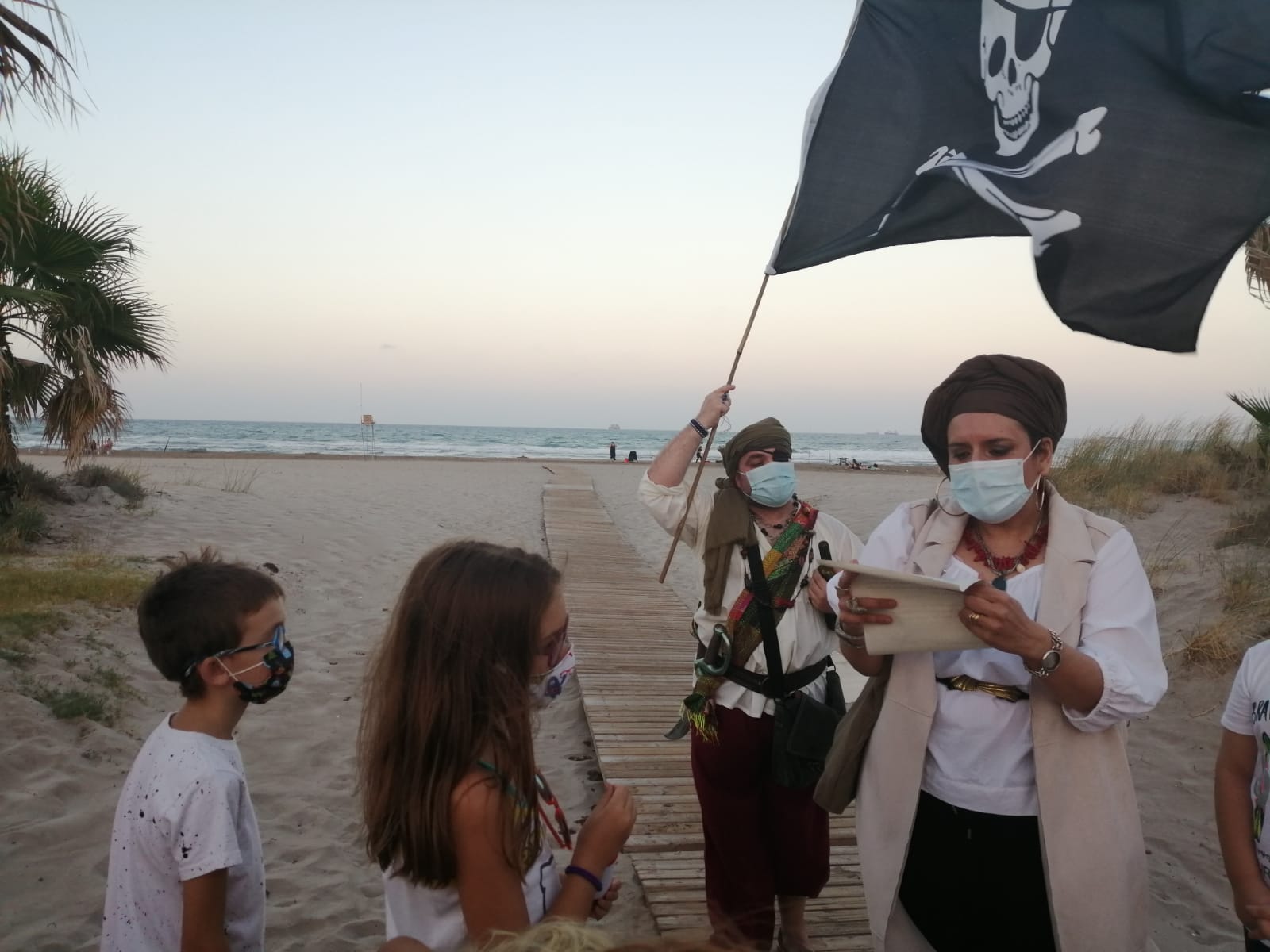 Dos personas vestidas de piratas teen una carta y enarbolan unabandera pirata en un paisaje de playa con dos niños mirando