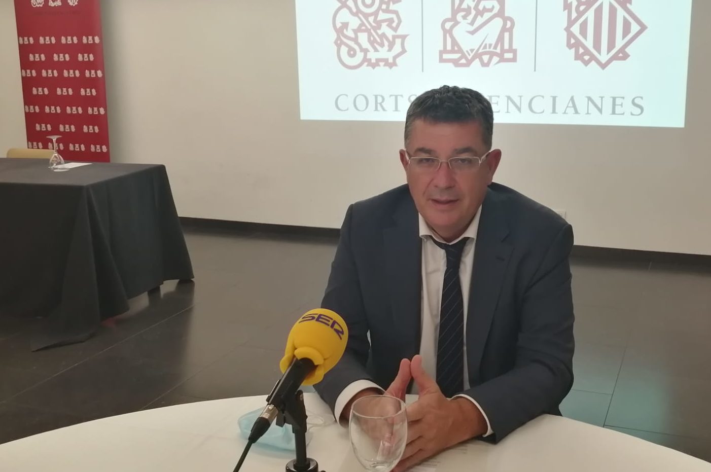 Imagen del president de les Corts Valencianes sentado con una ppantalal de proyección con el logotipo de los Corts tres él