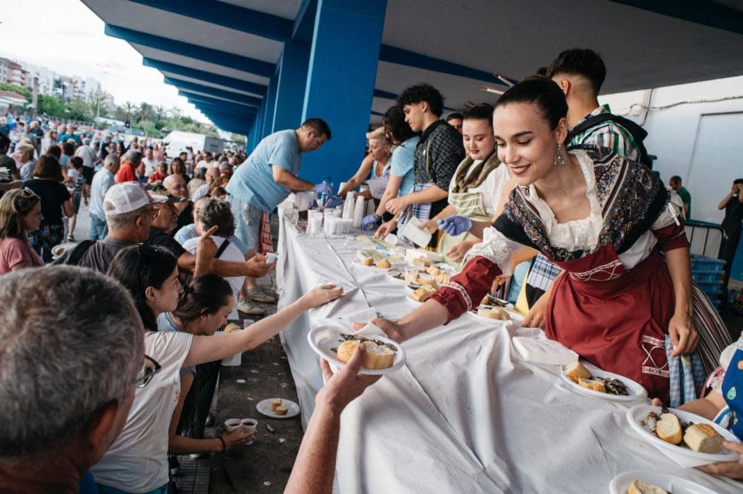 Imagen del reparto de la 'Torrà de la sardina' con festeras y festeros con indumentaria tradicional repartiendo platos a la gente desde la lonja