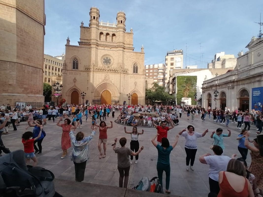 Imagen zenital de la plaza Mayor de Castellón con cients de personas repartidas y los brazos en alto bailando el ball perdut. Al fondo, la fachada d ela concatedral. Las personas van vestidas de calle y no con indumentaria tradicional