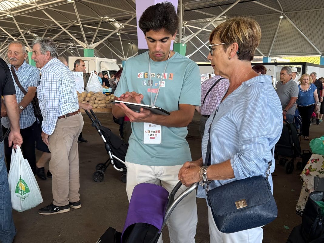 Un chico con una tablet y una identificación colgada al cuello habla con una mujer con un carro de la compra en el espacio del mercado de lunes de Castellón